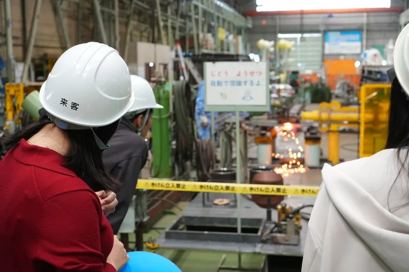 工場の中で、「来客」と書かれた白いヘルメットをかぶった見学者が、自動溶接の火花が散るところを柵の手前から見ている