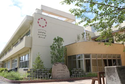 廃校になった小学校の校舎を使った建物。白い校舎の壁に「なめがたファーマーズヴィレッジ」の文字とロゴがあり、入口の前に「日本の農業をステキにしよう！」と彫られた石が置かれている