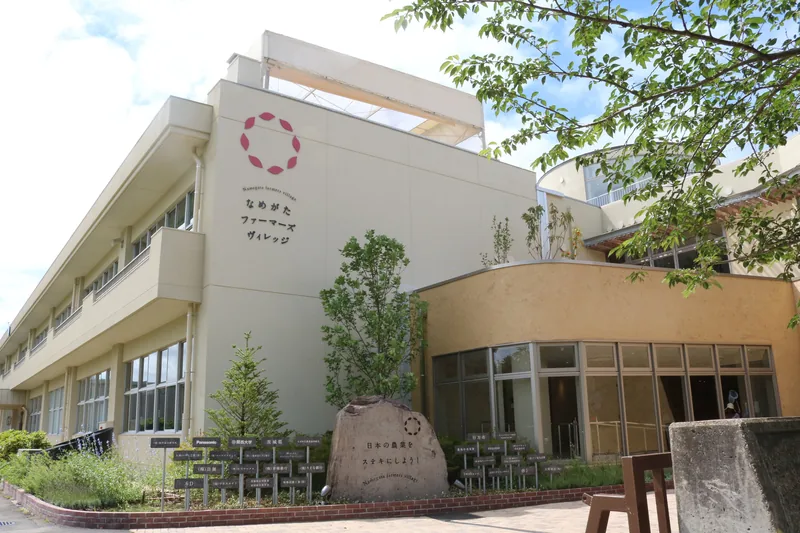 廃校になった小学校の校舎を使った建物。白い校舎の壁に「なめがたファーマーズヴィレッジ」の文字とロゴがあり、入口の前に「日本の農業をステキにしよう！」と彫られた石が置かれている